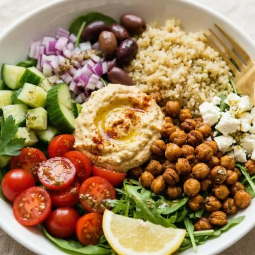 Overhead view of a Mediterranean chickpea bowl with golden crispy chickpeas, hummus dollop, red onion, olives, cucumber, tomatoes and lemon wedge