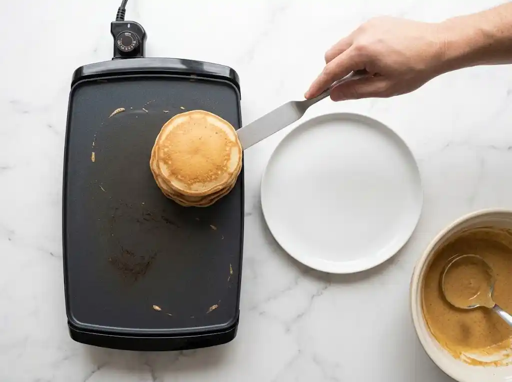 Transferring freshly cooked, warm pumpkin pancakes with pancake mixfrom an electric griddle onto a clean white serving plate.