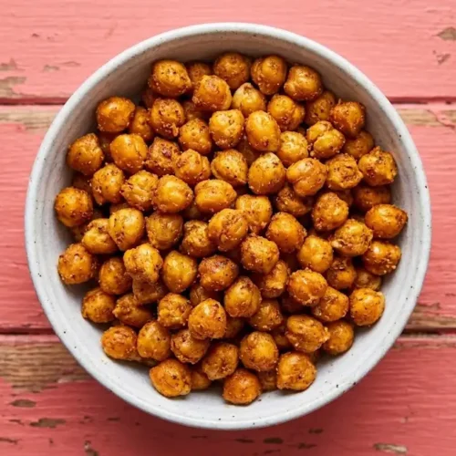 Top-down view of a bowl filled with crispy, golden air fryer chickpeas seasoned with spices on a rustic pink wooden table.