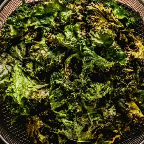 A close-up view of perfectly crispy air fryer kale chips inside a metal fryer basket against an orange tile background