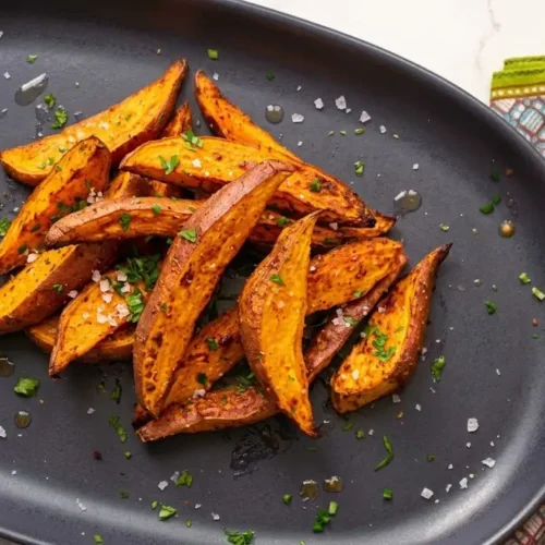 "Golden-brown roasted sweet potato wedges on a dark platter, seasoned with coarse sea salt and fresh chopped green herbs (parsley). Perfect healthy side dish or snack. Includes serving tongs."