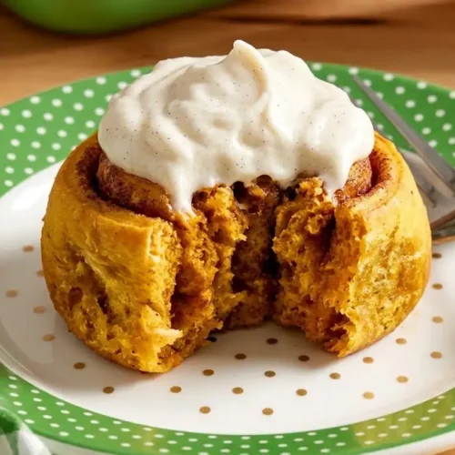 A single, baked pumpkin cinnamon roll covered in creamy cream cheese frosting on a white plate with a fork.