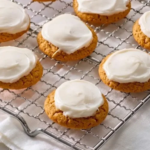 "A close-up photograph of several soft, cake-like pumpkin cookies topped with a generous swirl of creamy cream cheese frosting, resting on a wire cooling rack and ready to serve."