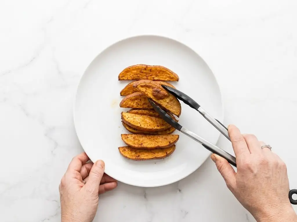 step4 Air Fryer Sweet Potato Wedges A person's hand uses tongs to carefully place a cooked sweet potato wedge onto a neat stack of roasted wedges on a white round plate. Elegant plating for a healthy side dish.
